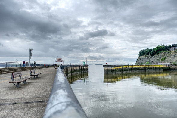 Looking along a railing on the barrage outer harbour arm towards the entrance to the lock complex. In the distance beyond the entrance, the islands of Flat Holm and Steep Holm are visible and the cliffs of Penarth Head are on the right. Fishermen line the left hand harbour wall.