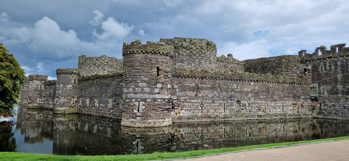 Beaumaris castle, surrounded by its moat, in which you can see its reflection. A dark cloudy sky is above.