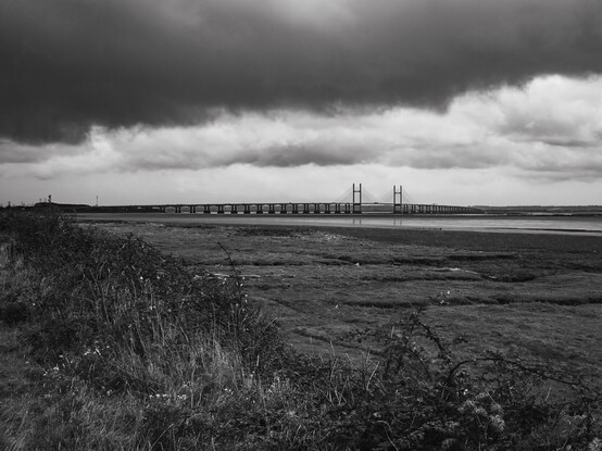 The Second Severn Crossing:

A black and white photo of this long bridge as it crosses the Severn. The tide is out revealing the substantial concrete pillars supporting most of the bridge, The middle section however, is supported by wires suspended from two tall towers. The towers are reflected in the water below the bridge. Above thick, dark clouds threaten rain