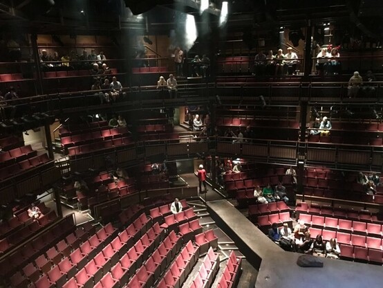 A theatre auditorium. The chairs are red, and many of them are empty as the audience comes in.