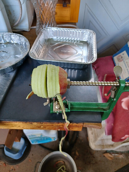 A ripe apple mounted on an apple peeler/corer/slicer device.  Device is painted green with silvery metal shaft and cutting pieces. Apple has been partially peeled and sliced.  Aluminum baking pans in background.  Apple peel is dangling down to a pot on the floor.