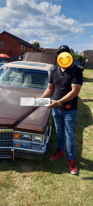 A man standing next to a brown chevrolet holding a sketchbook with a sketch of the chevrolet.