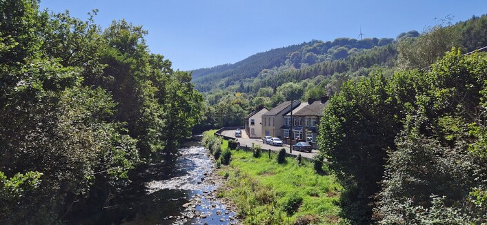 A view of the village from the river bridge. Trees of various types and shades of dark green flank both sides of the river. A bright green grassy banking is to the right with a little row of houses beyond. In the distance the mountain looks hazey because of the heat and humidity. A clear blue sky is above.