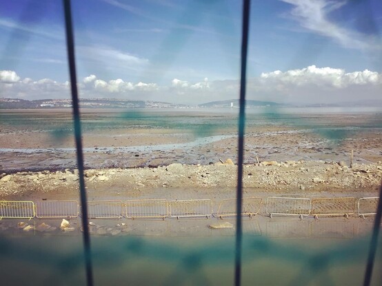 Looking across Swansea bay from Mumbles, low tide - mud, silt, stone bedrock, sea, industry, pipes, piers, rusted remnants, and pipes. As people still flock in sun to walk it’s open shores and open blue skies.