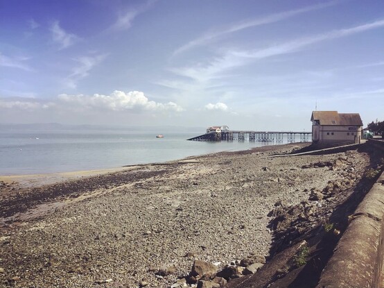 Looking across Swansea bay from Mumbles, low tide - mud, silt, stone bedrock, sea, industry, pipes, piers, rusted remnants, and pipes. As people still flock in sun to walk it’s open shores and open blue skies.