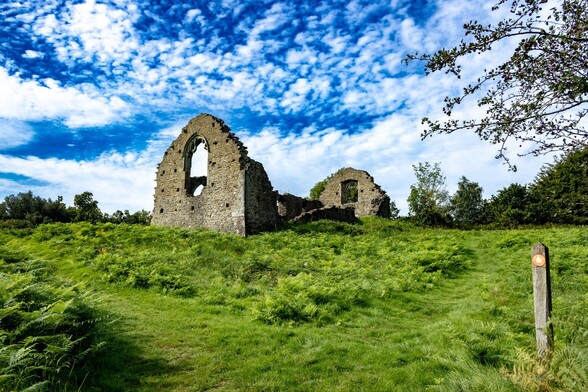 Capel Mair:

Looking up a grass and bracken covered slope towards the Eastern end of a ruined grey stone chapel. Both arched end walls are largely intact, with some stone details around the large windows still visible. The side walls are more damaged, with the closer one to the camera mostly fallen down. The sky is a deep blue with a high thin covering of white cloud breaking up