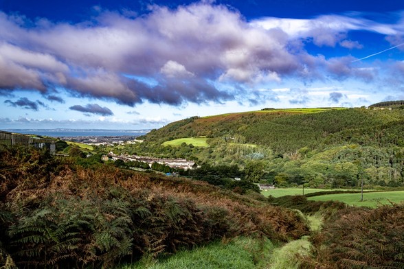 Looking down the Afan Valley, over Aberafan and Swansea Bay to Swansea:

Looking down a, mostly, bracken covered hillside to a V shaped valley, the opposite side of the valley is mostly wooded with  some farmland on top. A  village of brown roofed, white houses sits in the valley. Further on the valley opens out to a tightly packed town, which in turn gives way to a blue strip of sea. In the far distance the coastline and buildings of Swansea can be seen over the bay. Above, fluffy white clouds are liberally spread over a deep blue sky.