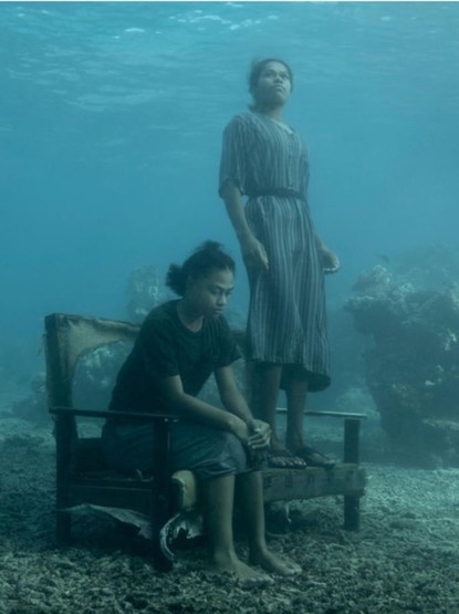 Two Fuji women one sits on a bench looking down, expressionless. The other standing on the bench, looking skyward expressionless. The scene is underwater, muted tones of aqua blues