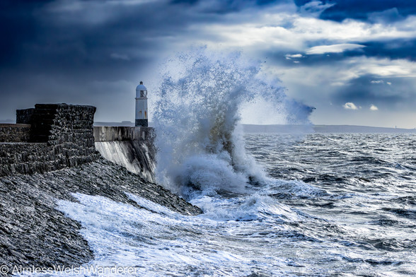 Stormy Seas at Porthcawl Lighthouse:

A grey, stone harbour wall leads out into rough, grey seas. A small white lighthouse stands at it's end. A large wave breaks at the the base of the harbour wall, sending spray high above the lighthouse. In the distance, the cliffs of the Glamorgan Heritage Coast can be seen under stormy grey skies