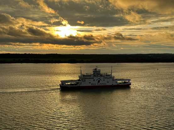 Sunset across Southampton water. Calm sea with a small ferry sailing up the water towards Southampton docks. A silhouette of the land on the far side of the water is in the distance with orange skies reflected in the water at sunset. Mixed clouds hand over the water with a yellow tint from the sun.