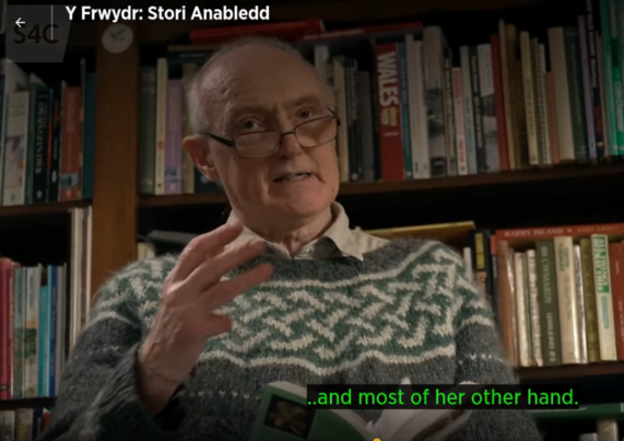 An old man with glasses gesticulating; behind him are full bookshelves