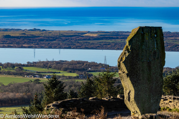 View from The Bro Stone:

A large, irregular shaped stone stands vertically, the word "bro" can just be seen carved into it's rear face. A low stone wall forms a circle around it and the ground drops sharply away in front of it with the tops of conifer trees visible. further ahead, and below, green farmland is punctured by patches of leafless woodland and bounded by the M4 motorway, where a few vehicles can be seen. There is then a large rectangular reservoir, it's still water reflecting the image of electricity pylons.  Further on a band of rough shrub-land leads to grass covered sand dunes at the edge of a still blue sea
