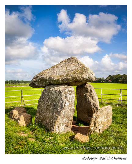 Photo of a small chambered tomb or dolmen with a pyramidal capstone resting on three upright stones, surrounded by a metal fence and standing in a grassy field.
