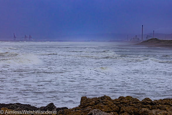 Stormy Sker Beach:

Looking over rough seas of waves and white surf to the cranes and chimney stacks of TATA steel in Port Talbot. The sky is dark and brooding and a gloomy haze covers the distance hills
