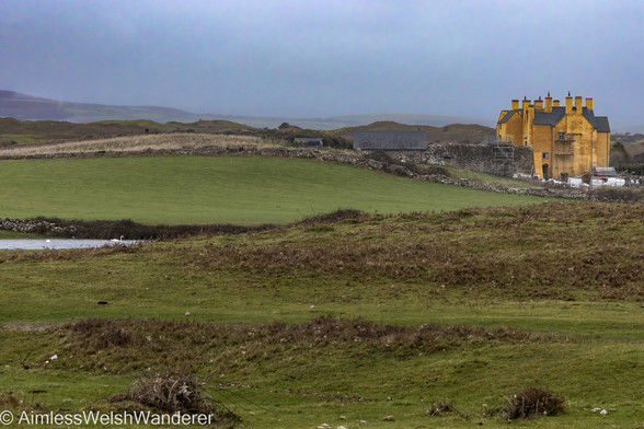 Sker House:

Looking over undulating, rough grass fields seperated from one another by old dry stone walls. The recent rain has formed a pond at the edge of the nearest field where two white, Mute Swans swim serenely. In the middle distance a large imposing house stands. It has a dark slate roof punctuated by numerous chimneys (9 can be seen) and is rendered in a bright, mustard yellow colour. A tall grey stone wall runs away from the house and scaffolding rests against both the wall and the end of the house. The sky is a stormy dark grey
