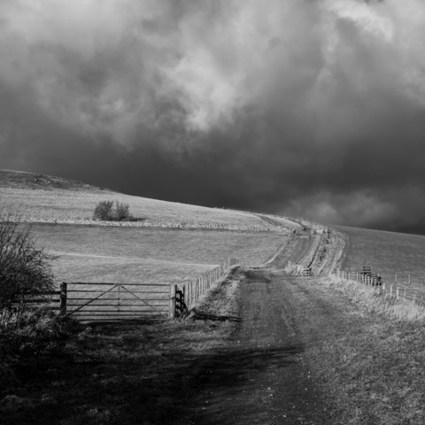 A black and white photograph. A track leads up and around a hill, with a gate in the foreground and fields beyond. The sky is dark and apocalyptic. 