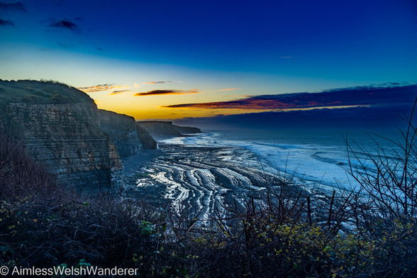 Sunrise over the Glamorgan Heritage Coast:

Looking along a stretch of coastline from a high vantage point. There is a long beach with sections of rock and sand, all of it backed by tall, sheer cliffs in which narrow layers of different rock types can be seen. In the middle distance, a waterfall flows onto the beach from the cliffs. The sun is rising from behind the cliffs, turning the sky golden and a bank of clouds to burn with red fire. A single contrail can be seen against a clear patch of dark blue sky