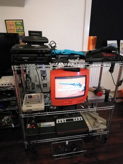 Wireframe shelving homing multiple retrogaming consoles, several with lightguns attached. They are centred around a bright orange 14" CRT TV.