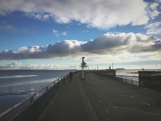 A landscape picture of Cardiff bay barrage. Look across low tide mud flats and the barrage reaching out like a road. Distant theatrical clouds, a single figures walks out. Low winter sun.
