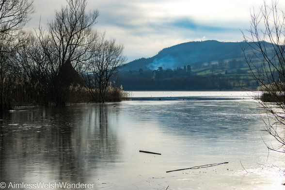 Frozen Crannog:

Looking out at Llangorse lake, which has frozen at the edges, in the middle distance the silhouette of the cone shaped roof of the crannog can be seen nestled amongst reeds and trees. In the distance smoke rises from a wooded area on the far bank before the land rises gently to a field covered hill 