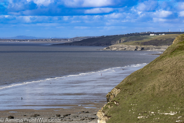 Traeth Mawr:

Looking along a long sandy beach from a high vantage point. The beach curves around to the right disappearing behind the high cliffs. In the distance the buildings of Southerndown, and further on, Porthcawl, can be seen across a calm, dark grey sea. Above, thick clouds are building in an otherwise blue sky