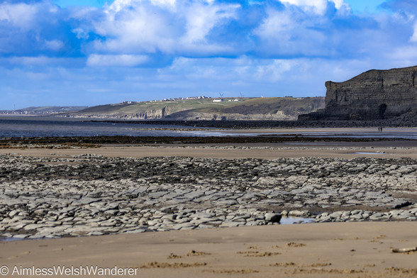 Monknash Beach:

Looking along a long beach of golden sand and mussel covered rocks. In the middle distance, the beach curves around to the right disappearing behind a high rocky promontory. In the far distance, across a calm, dark grey sea, the houses of Southerndown can be seen sitting high above the water. Above, thick clouds are building in an otherwise blue sky