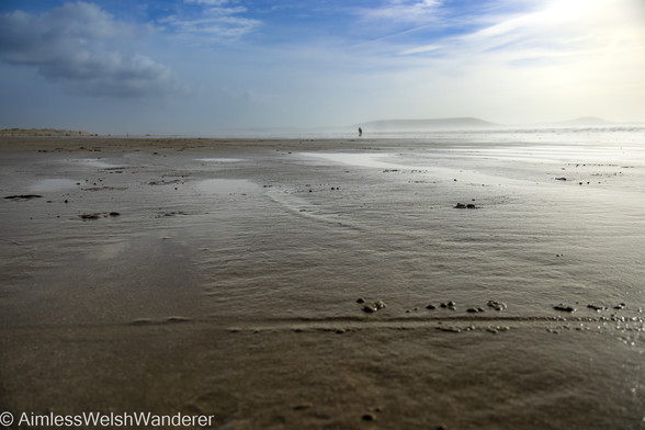 Looking along a flat, wet sandy beach with the camera positioned close to the ground. Sand dunes can be seen in the distance, to the left of the beach with the sea, overexposed by a bright sun is breaking on the right. In the distance, over the water, the shadows of the Gower peninsular emerge from a sea haze. Above, blue sky is showing through white clouds.