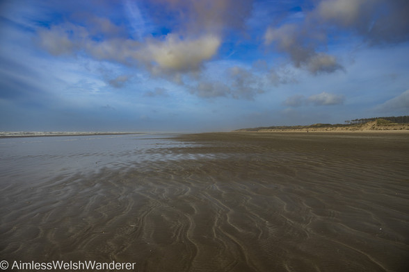 Looking along a flat, wet sandy beach rippled with the memory of recent waves. Sand dunes can be seen backing the beach on the right, while the sea is breaking on the left. Above, a few white clouds stand against a blue sky.