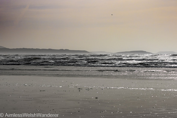 Looking out to sea from a flat sandy beach as low rolling waves sparkle in a bright sun. In the distance, the Gower peninsular emerges from a hazy sky, tinged orange by a bright sun