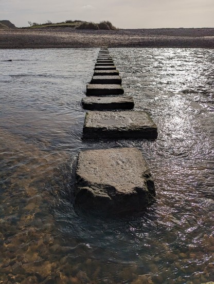 Stepping stones across a fast flowing river with light reflecting on it, and a sandy dune on the other side.