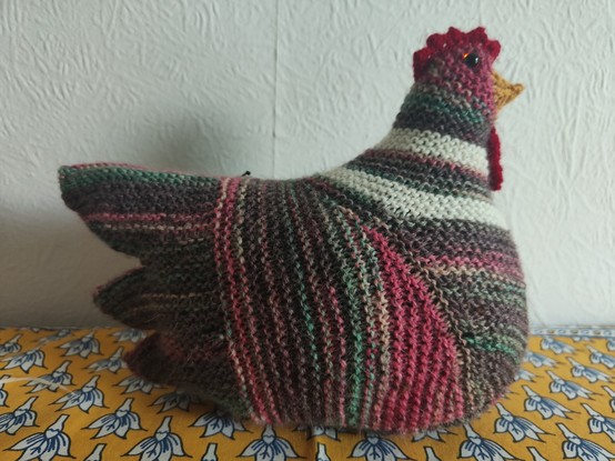A knitted chicken. It is various colours - mostly brown, pinky red, some green and with cream stripes.