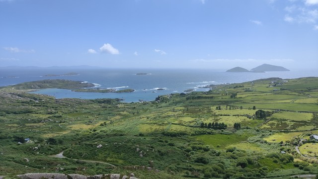 Looking down from a viewing point beside a road down over fields towards the coast line. It's a fine sunny day with blue sky and some small clouds. The fields are of various shapes and sizes, and are many shades of green, with the clouds throwing shadows. The sea is quite blue, though where it meets the land it is making lots of white breakwater. On the horizon on the right hand side are two sizeable islands.