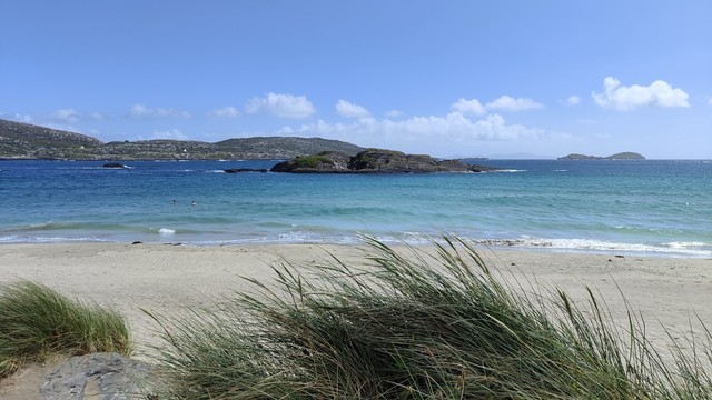 A beach in a small somewhat sheltered bay. The sky is blue with white clouds, and the water appears to be various shades of deep blue and turquoise. There's a sandy beach, with some marram grass in the very bottom of the picture.