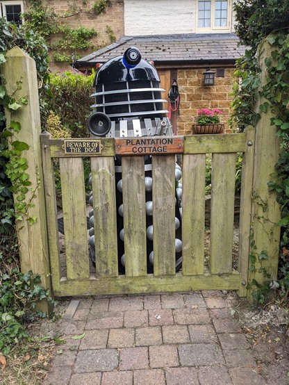 A Dalek behind a cottage gate with a sign on it saying beware of the dog. 