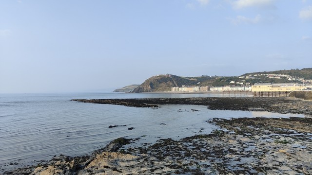 Looking over rocks and the sea towards the promenade of a Victorian seaside town. In the distance there is a row of buildings, with green hills behind. It's a hazy, late summer evening, with pale blue skies and the odd cloud visible.
