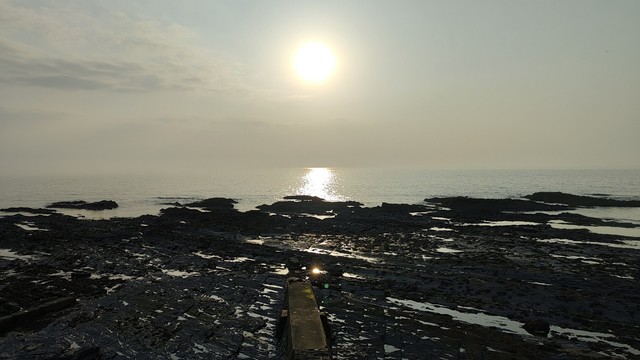 Looking out over rocks and small pools of seawater, towards the sea and the sun, which is low in a hazy sky.