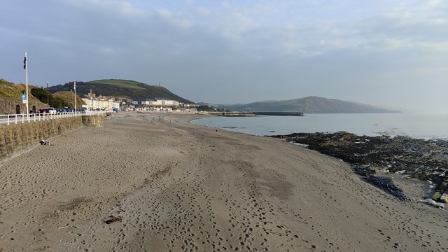 Looking down on to the beach, where there's many sets of footprints which go in many directions. In the distance there's various buildings, with hills beyond. It's a hazy evening, with clouds in the sky.