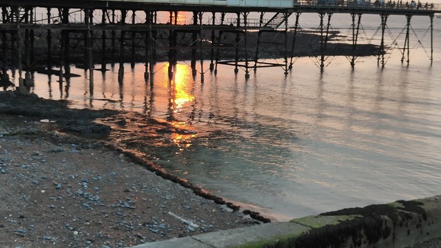 A section of beach and the sea, with the legs of the pier visible. It's close to sunset, and a glowing reflection of the sun is visible on the water.