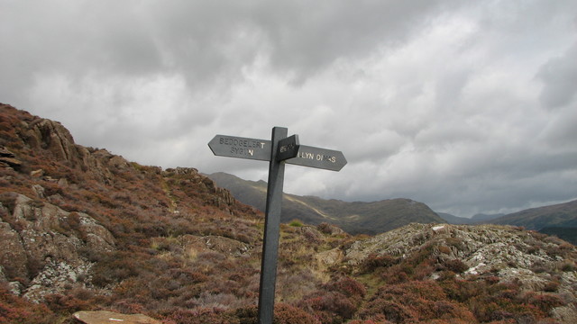 Photo of a remote signpost high above Beddgelert in the mountains. the sign post has 3 fingers set in rocky terrain with a grey moody sky.