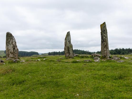 Photograph of the 3 Glengor Standing Stones, set in a green field, with hills and woods in the background.