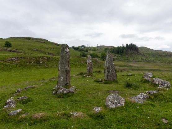 Photograph of the 3 Glengor Standing Stones, set in a green field, with hills and woods in the background.