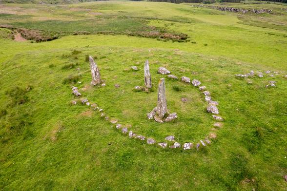 Drone photograph of the 3 Glengor Standing Stones, set in a green field, with hills and woods in the background. This photo also shows the ring of small stones surrounding the 3 standing stones