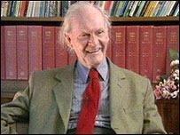 Colour photo of elderly balding man, seated in front of full bookshelves, laughing happily