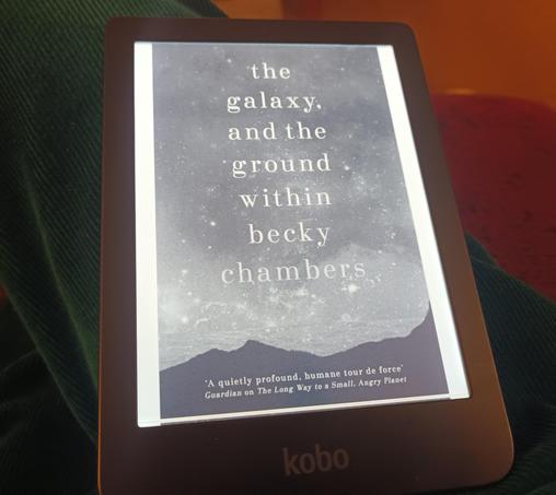 My ereader showing the cover of Becky Chambers's novel, The Galaxy and the Ground Within. It features the title and an image of a starry sky above a landscape.  