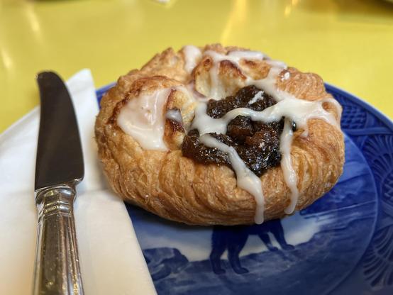 A close-up of a pastry topped with icing and a dark filling, served on a blue plate alongside a knife and napkin, placed on a yellow table.