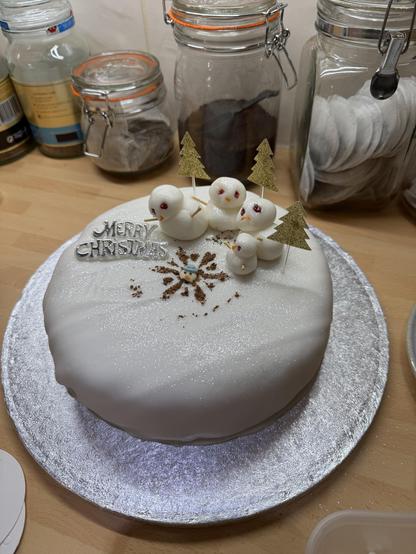 Christmas cake with flat white icing. 4 snowman with red eyes stand around a chocolate star formation on the ground. The decapitated head of an ordinary snowman lies in the middle of the star. The sign says Merry Christmas 