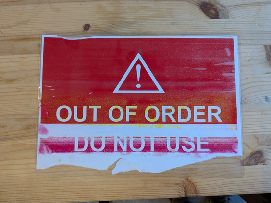 a laminated sheet of paper, which says "out of order, do not use" against a red background. the paper has white streaks, crumples, and a tear along the bottom from getting jammed while printing.