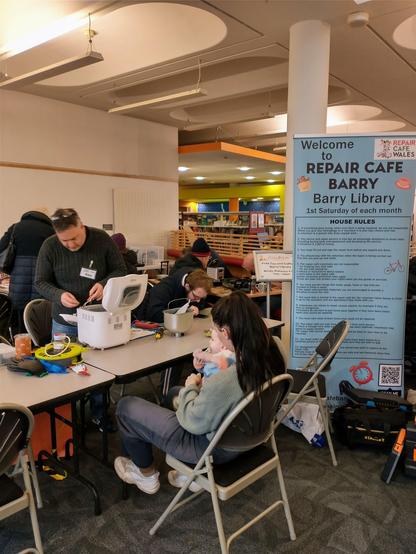 View into Barry repair cafe. To the right is a large blue sign reading "welcone to repair cafe Barry, Barry library" with house rules on it. To the right are tables laid out with chairs. In the foreground a person with light skin tone and long dark hair sits with a baby on their lap while Alex, a man with light skin tone and short dark grey hair stands behind the table fixing a bread maker. Behind him are more people sitting and standing.