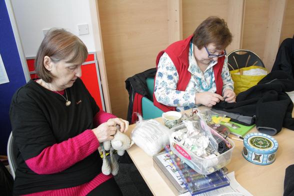 Ann, a woman with light skin tone and short brown hair, sewing a black jacket while Juliet, a woman with light skin tone and shoulder length brown hair, sits beside her stuffing a small child's teddy. On the table in front of them are all sorts of sewing paraphernalia.