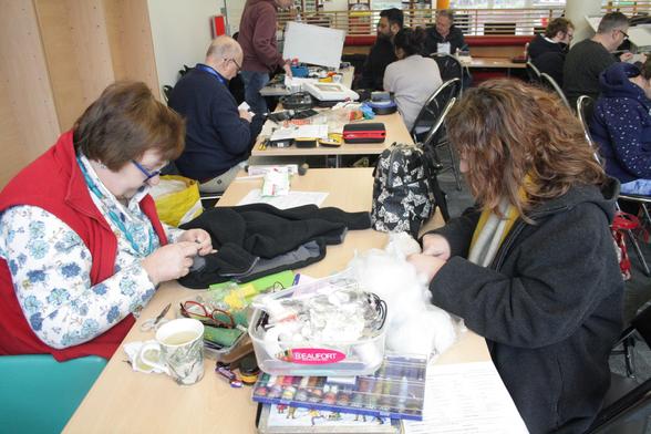 Ann, a woman with light skin tone and short brown hair, sewing a black jacket while across from her Mim, a woman with light skin tone and long curly brown hair, stuffs a small child's teddy. On the table in front of them are all sorts of sewing paraphernalia. Beind them are more tables of repair cafe with people chatting and working on things.
